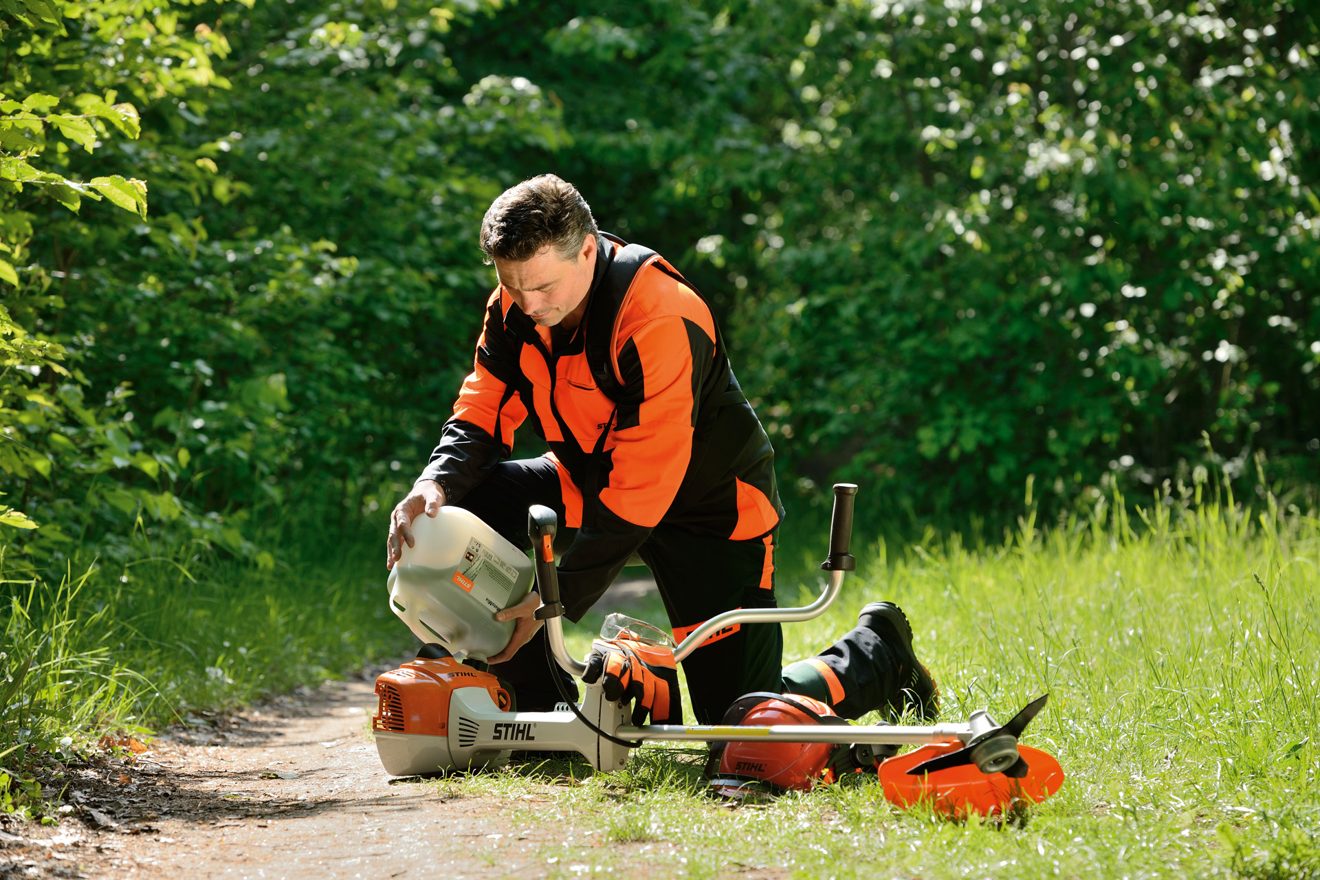 Un hombre llena el depósito de combustible de una desbrozadora STIHL con una garrafa con arbustos detrás de él