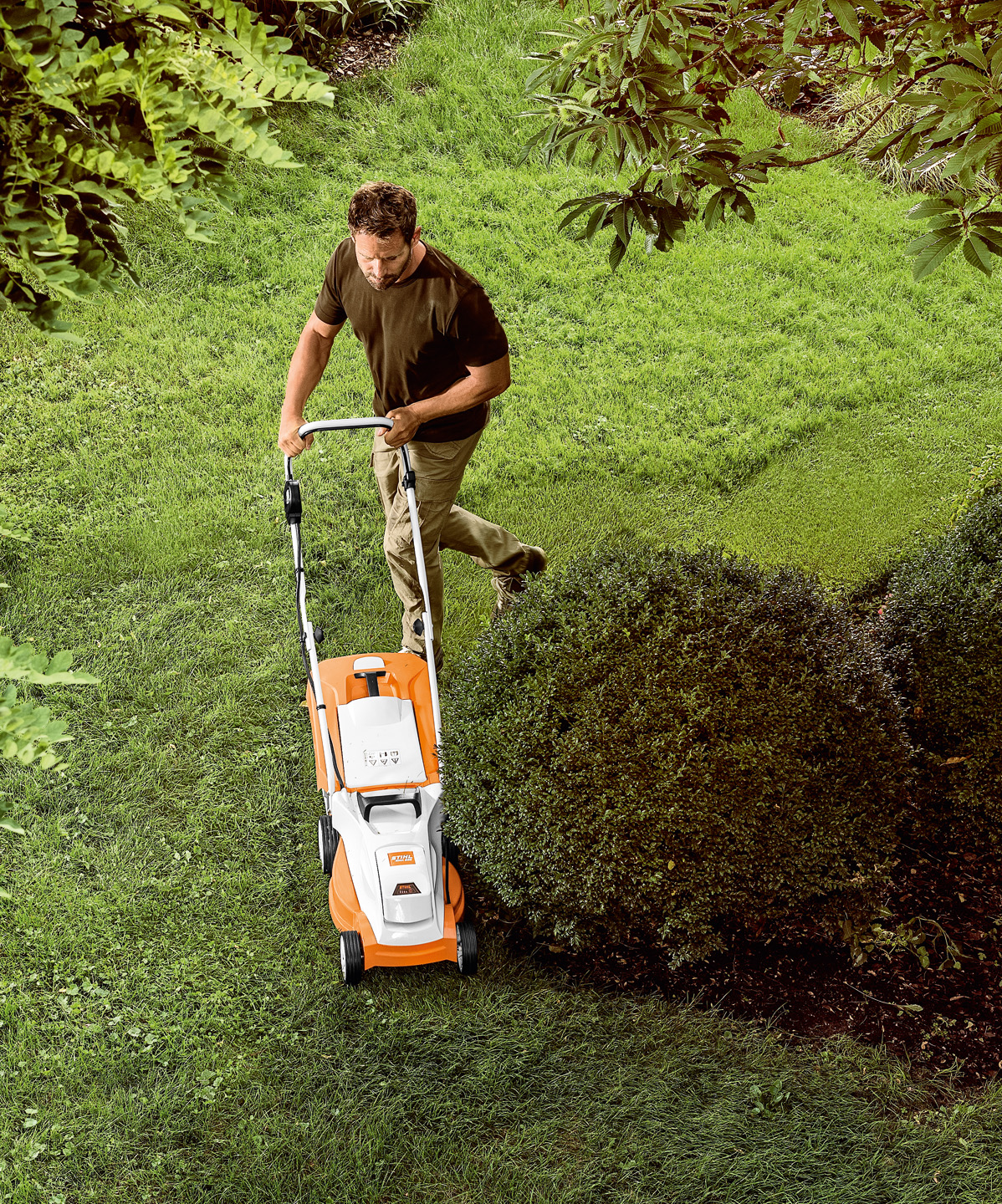 Vista desde arriba: Un hombre corta el césped con el cortacésped de batería STIHL RMA 235 alrededor de un pequeño árbol.